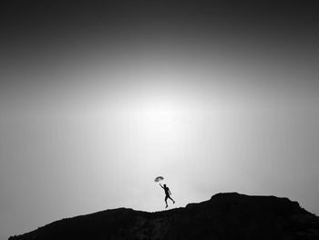 Silhouette person with umbrella jumping on cliff against sky