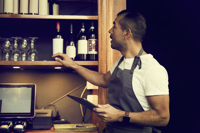 Male owner with digital tablet looking at wine bottles while standing in cafe