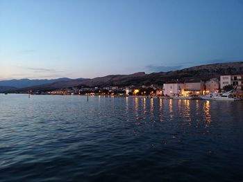 Illuminated buildings by sea against sky at dusk