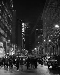 People on illuminated street amidst buildings in city at night against city skyline