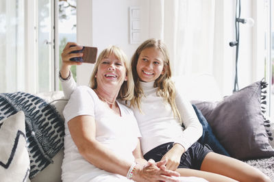 Smiling granddaughter taking selfie with grandmother through mobile phone on sofa at home