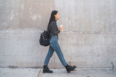 Side view of woman holding coffee cup walking by wall outdoors