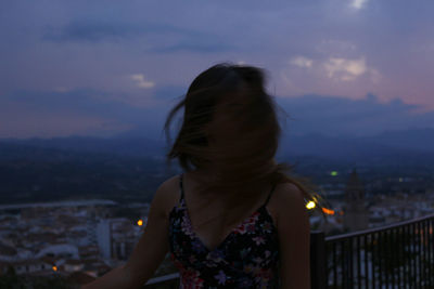 Woman with tousled hair standing by railing against cityscape at dusk