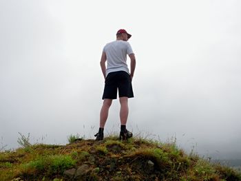 Man in cloud of fog. hiker in white black sportswear and red cap stand on rocky peak . around mist