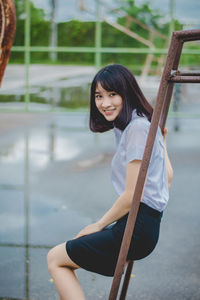 Portrait of smiling young woman sitting on metallic outdoor play equipment at playground during rainy season