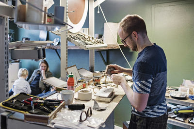 Male owner working at desk in eyewear workshop while family sitting in background