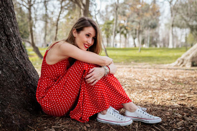 Side view of smiling woman looking away while sitting by tree trunk in park