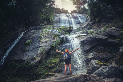Rear view of woman with arms raised standing on wood in forest against waterfall