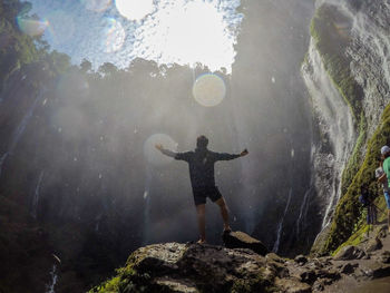 Full length of male hiker with arms outstretched standing against waterfall