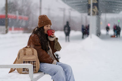 Young traveler girl drink hot tea at train station wait for transport departure for winter vacation