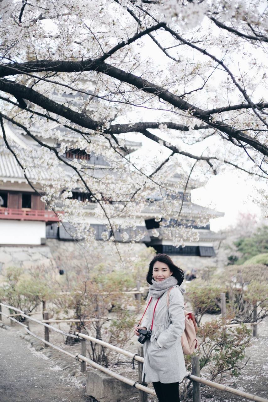 tree, one person, plant, young adult, standing, lifestyles, day, three quarter length, real people, leisure activity, nature, front view, architecture, adult, casual clothing, growth, women, focus on foreground, outdoors, cherry blossom, springtime, cherry tree