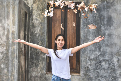 Portrait of smiling young woman throwing leaves while standing against wall