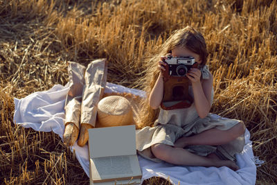 Girl child in a dress sitting on a mown field with a camera on a blanket with bread and a book