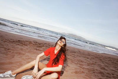 Young woman sitting at beach against sky