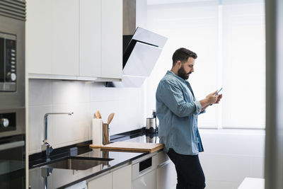 Man cooking crepes in the kitchen with a mobile phone in a denim shirt