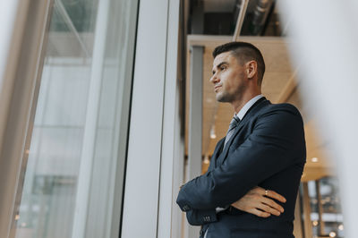 Thoughtful businessman standing with arms crossed looking out through window at office