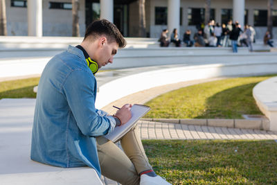 Side view of thoughtful smart male student in bright headphones studying in university square and writing in note book sitting on fence with crossed leg