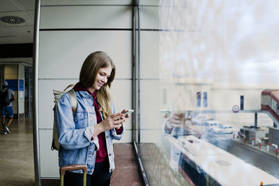 Young woman with suitcase using phone by glass window at airport