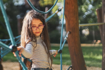 Portrait of a beautiful caucasian girl stands half-side on a rope swing in a park on a playground
