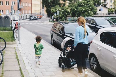 Mother pushing baby stroller while walking with son on sidewalk at city