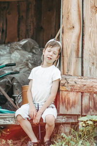 A tired boy sits leaning against the barn door with his eyes closed. 