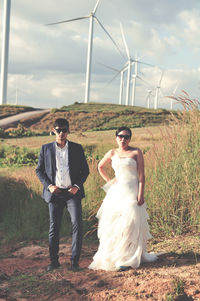 Portrait of bride and groom wearing sunglasses while standing on field against windmills