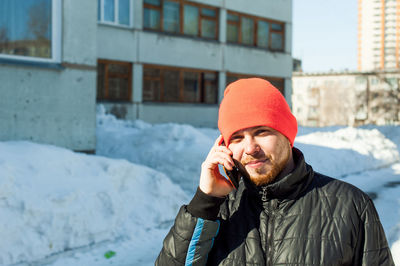 Portrait of man in warm clothing talking on mobile phone against building