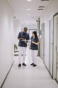 Full length of male doctor discussing over report with female colleague while standing in corridor at hospital