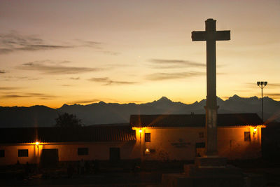 Silhouette cross by building against sky during sunset