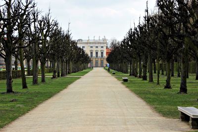 Way towards building flanked by bare trees against cloudy sky