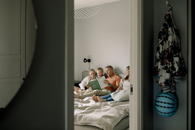 Man reading picture book for children while sitting on bed seen through doorway at home