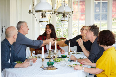 Happy mature friends having lunch together in dining room at home during social gathering