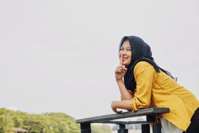 Young woman sitting on bench against clear sky