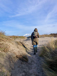 Rear view of a woman walking up a dune, while at the horizon a plume of smoke rises in the blue sky