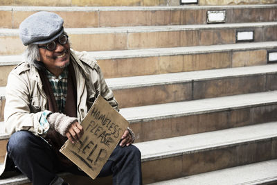 Smiling beggar holding placard with text while sitting on steps in city