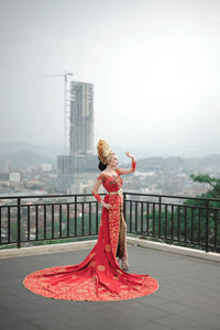 Woman with traditional custom wedding of indonesia standing by railing against sky in city