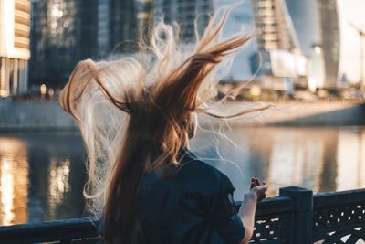 Rear view of woman with tousled hair standing against river in city