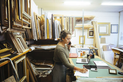 Confident young craftswoman talking on smart phone while writing in paper at workbench in framing store