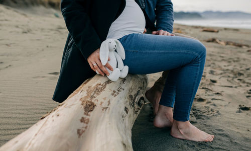 Low section of pregnant woman sitting with man on driftwood at beach