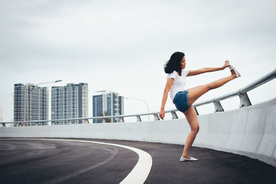 Full length side view of woman exercising by railing on bridge in city against sky