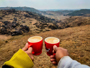 Cropped hand of couple holding coffee cup against landscape