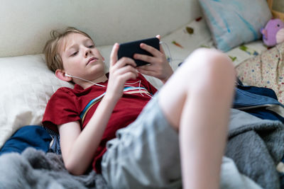 Young boy lying on bed looking at smartphone with earbuds in ears