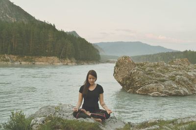 Young woman meditating at riverbank against sky