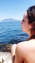 Rear of beautiful woman looking away while sitting by sea against clear sky during sunny day