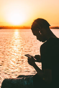 Side view of young man sitting at ocean using mobile phone against clear sky