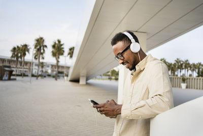 Man using mobile phone wearing headphones listening to music leaning by wall