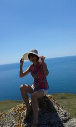 Woman holding sun hat while sitting on rock by sea against sky