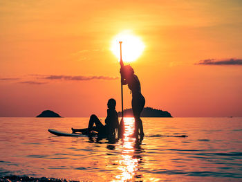 2 young women paddle boarding on sea against sunset