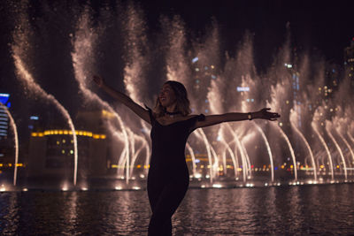 Woman with arms outstretched against fountain in city at night