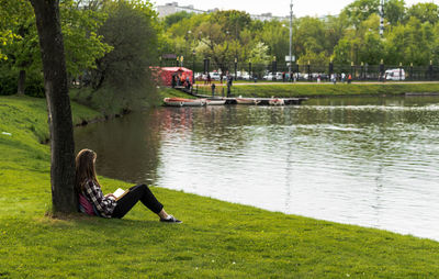 Woman sitting on grass by lake against trees